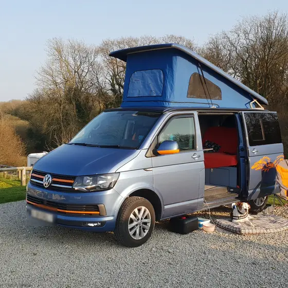 Furgoneta camper con el techo elevable y la puerta corredera abiertos en un camping, con la luz de la tarde