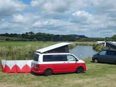 Furgoneta camper roja y blanca con techo elevable en un camping junto a un río